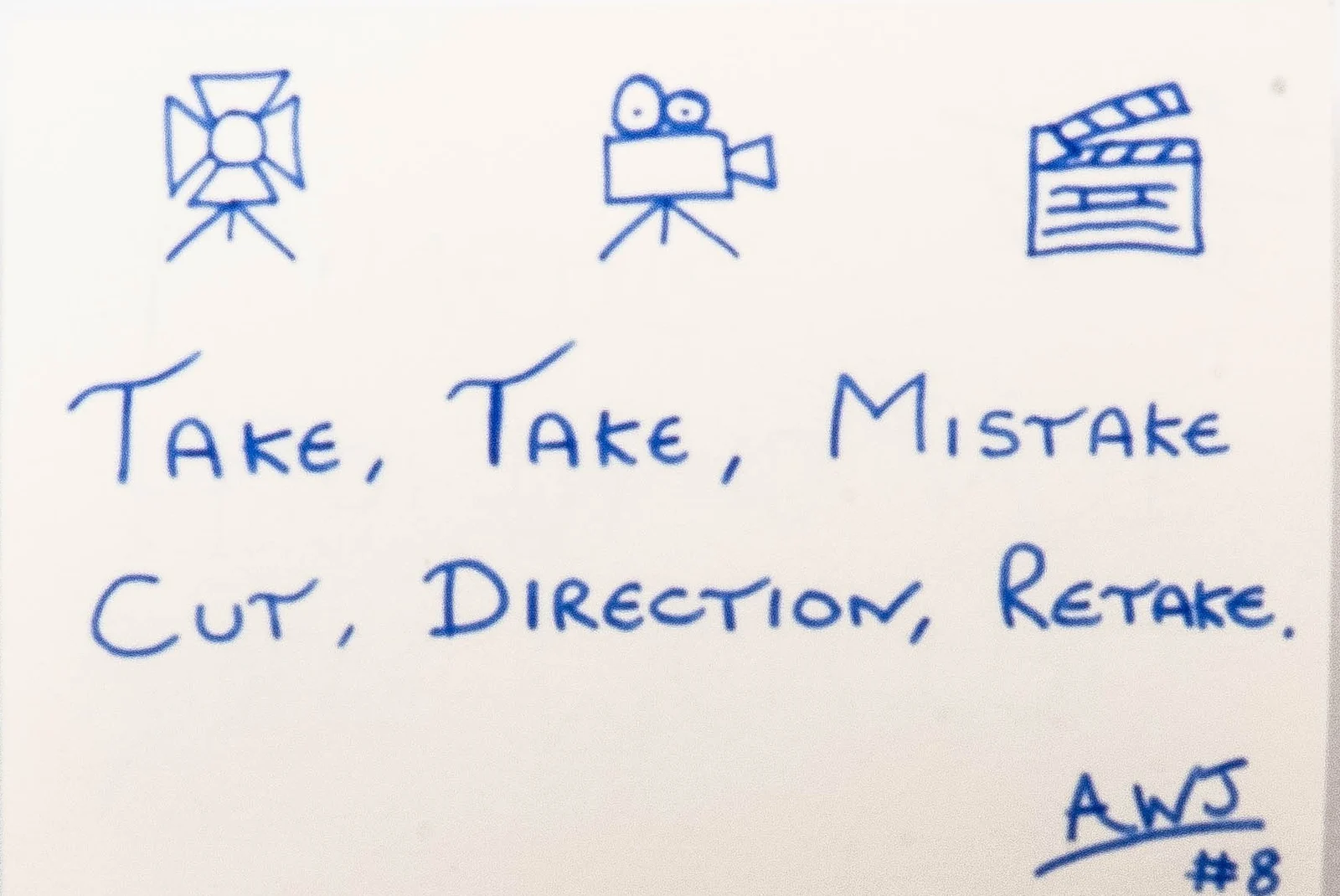 Handwritten in blue ink beneath three small drawings of a studio light, a camera and a clapperboard: Take, take, mistake. Cut, direction, retake. Signed AWJ, number 8.